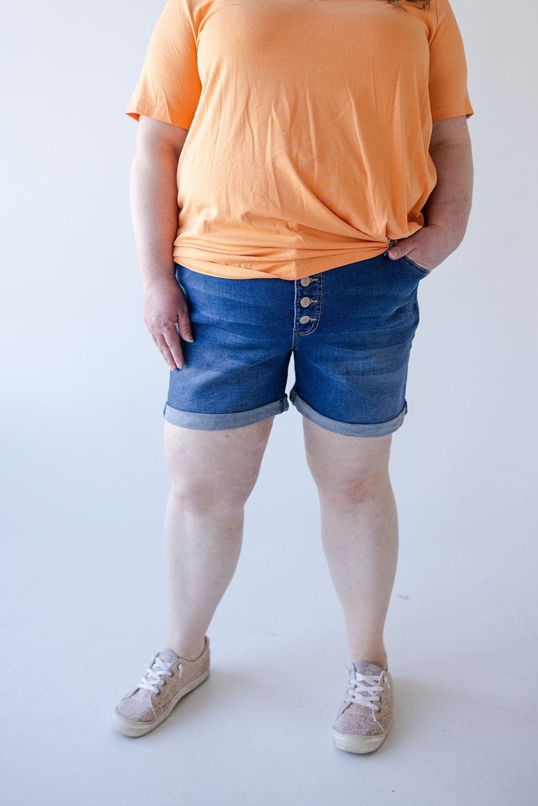 Person wearing an orange shirt, BUTTON FLY CUFFED SHORTS IN MEDIUM WASH by Love Marlow with 5 pocket construction, and beige sneakers, standing with one hand in pocket.