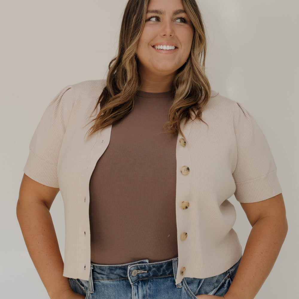 Woman wearing a beige cardigan over a brown top and blue jeans against a white background
