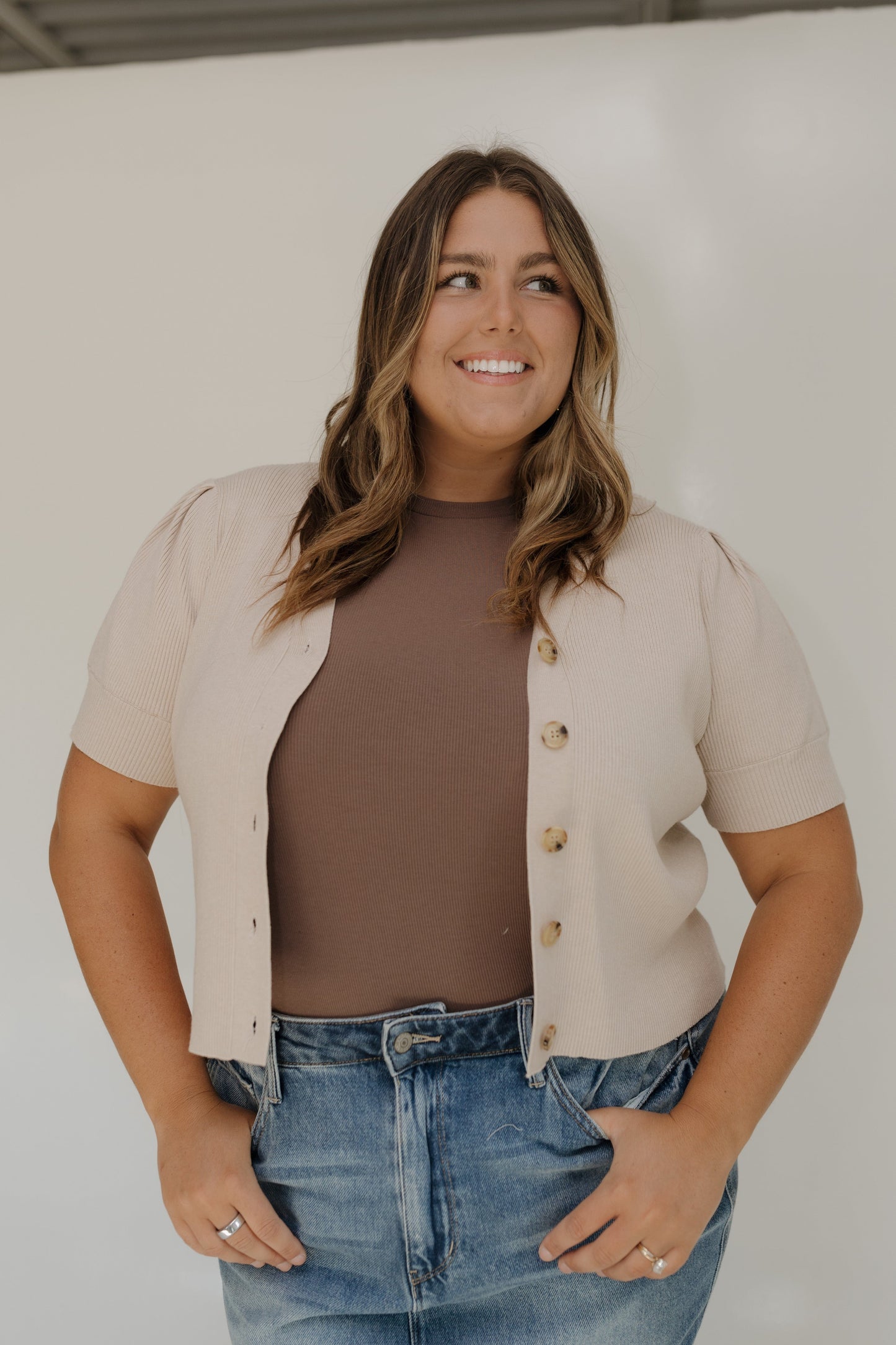 Woman wearing a beige cardigan over a brown top and blue jeans against a white background