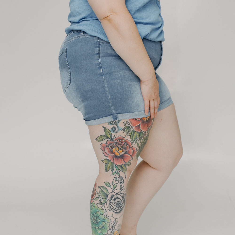 Person wearing a denim skirt and black sandals with floral tattoos on a plain background