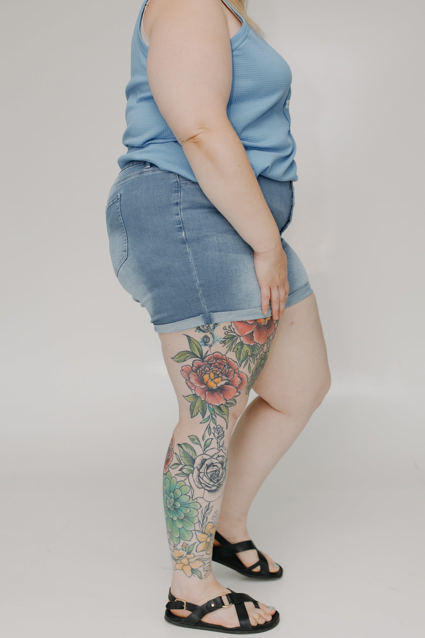 Person wearing a denim skirt and black sandals with floral tattoos on a plain background