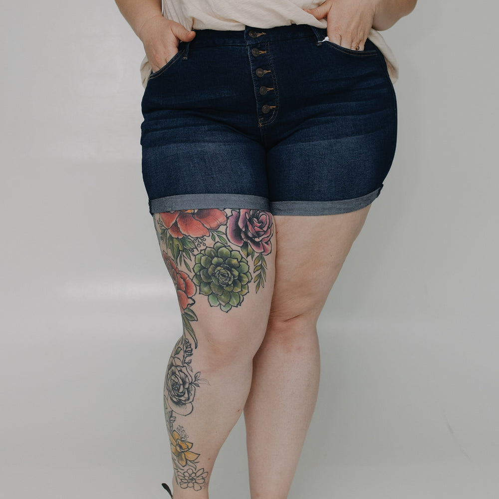 Person wearing dark denim shorts with floral tattoos on legs against a plain background