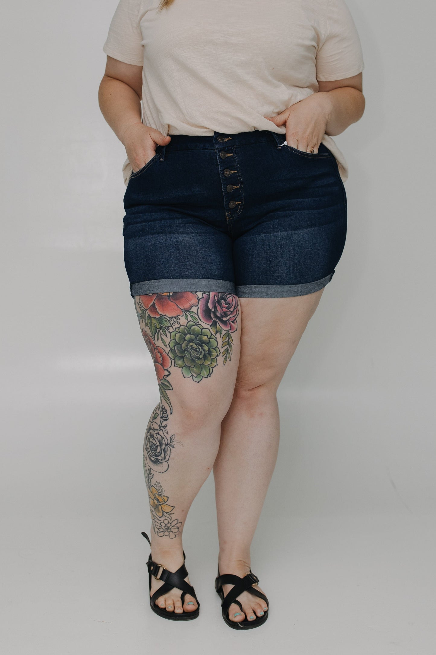Person wearing dark denim shorts with floral tattoos on legs against a plain background