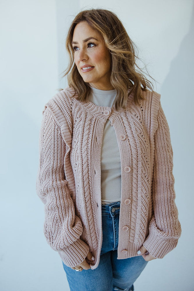 A person wearing a grey rose button-front cable knit cardigan by Yesta and jeans stands against a light background. They have shoulder-length hair and are looking to the side.