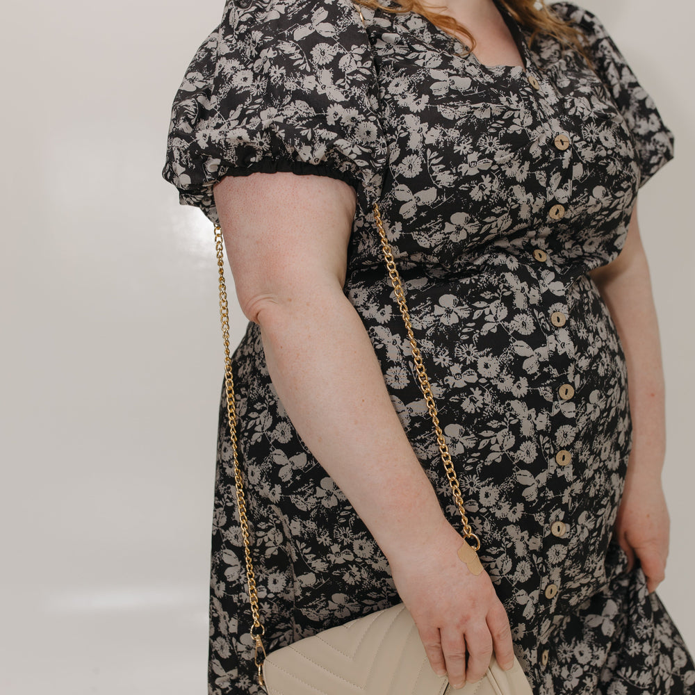 Woman wearing a black floral dress holding a beige handbag against a plain background