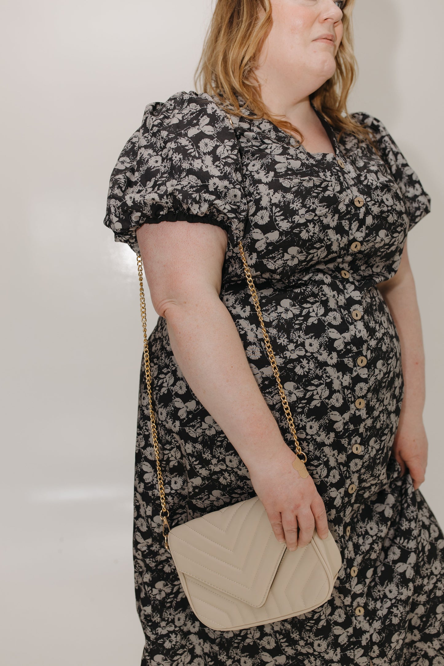 Woman wearing a black floral dress holding a beige handbag against a plain background