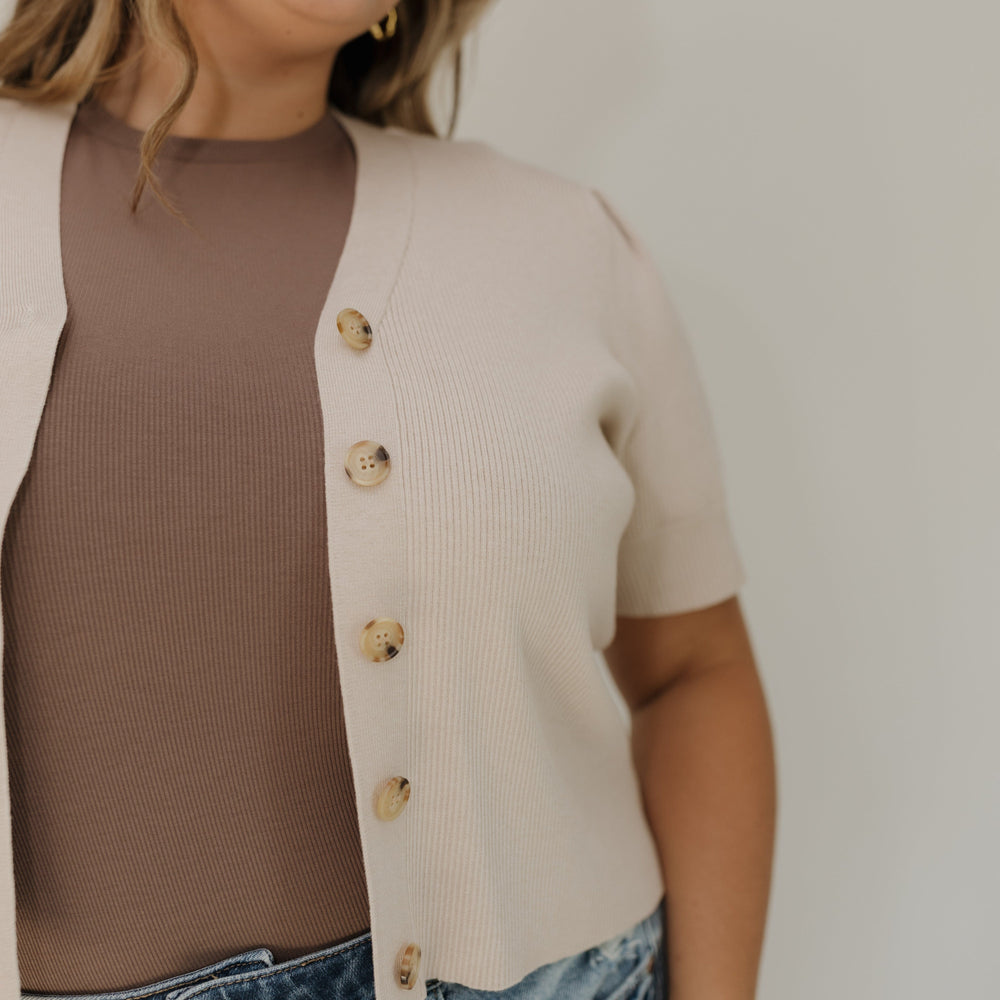 Woman wearing a beige cardigan over a brown top and blue jeans against a plain background
