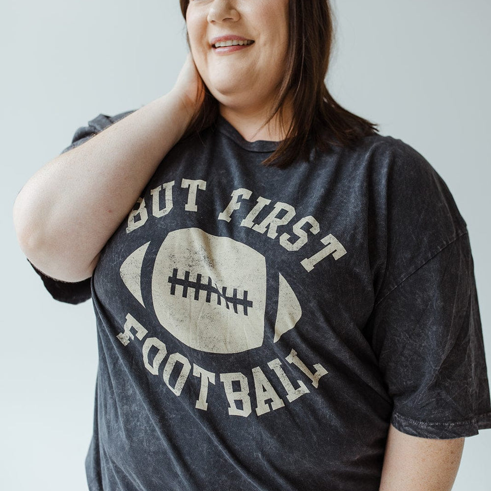 A person smiling while wearing a "But First Football" graphic tee from Love Marlow, made with super soft fabric and featuring a stylish mineral wash black finish.