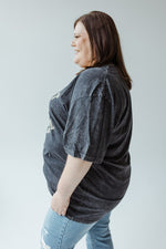 A person with long hair wearing the "BUT FIRST FOOTBALL" graphic tee by Love Marlow in mineral wash black and blue jeans stands facing away in a neutral-colored room.