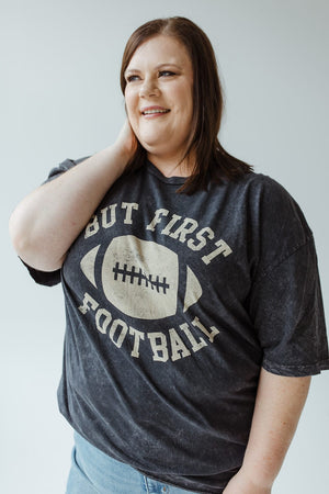 A person smiling while wearing a "But First Football" graphic tee from Love Marlow, made with super soft fabric and featuring a stylish mineral wash black finish.