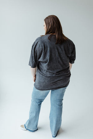 A person with long hair wearing the "BUT FIRST FOOTBALL" graphic tee by Love Marlow in mineral wash black and blue jeans stands facing away in a neutral-colored room.