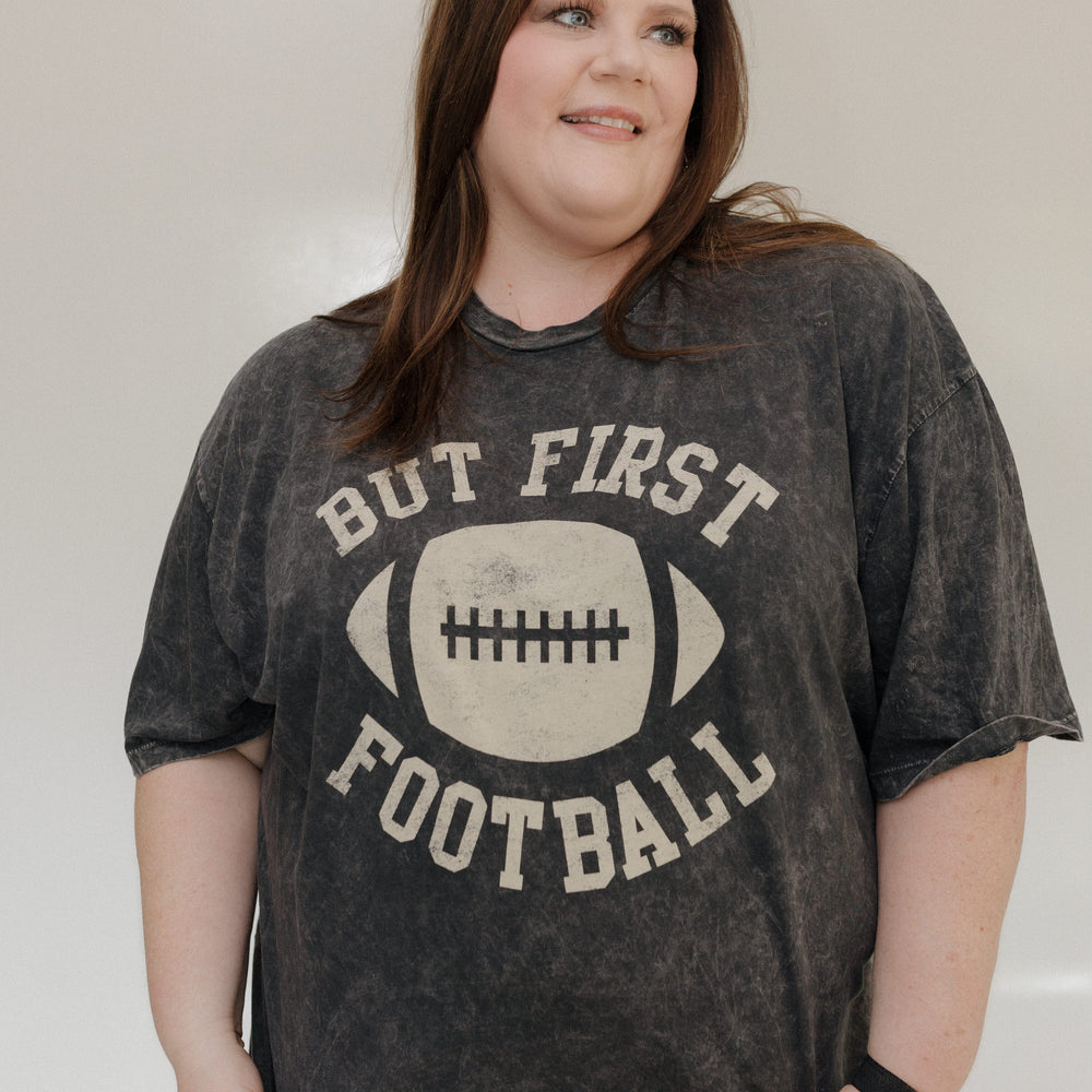 "BUT FIRST FOOTBALL" GRAPHIC TEE A person smiling while wearing a "But First Football" graphic tee from Love Marlow, made with super soft fabric and featuring a stylish mineral wash black finish. Love Marlow
