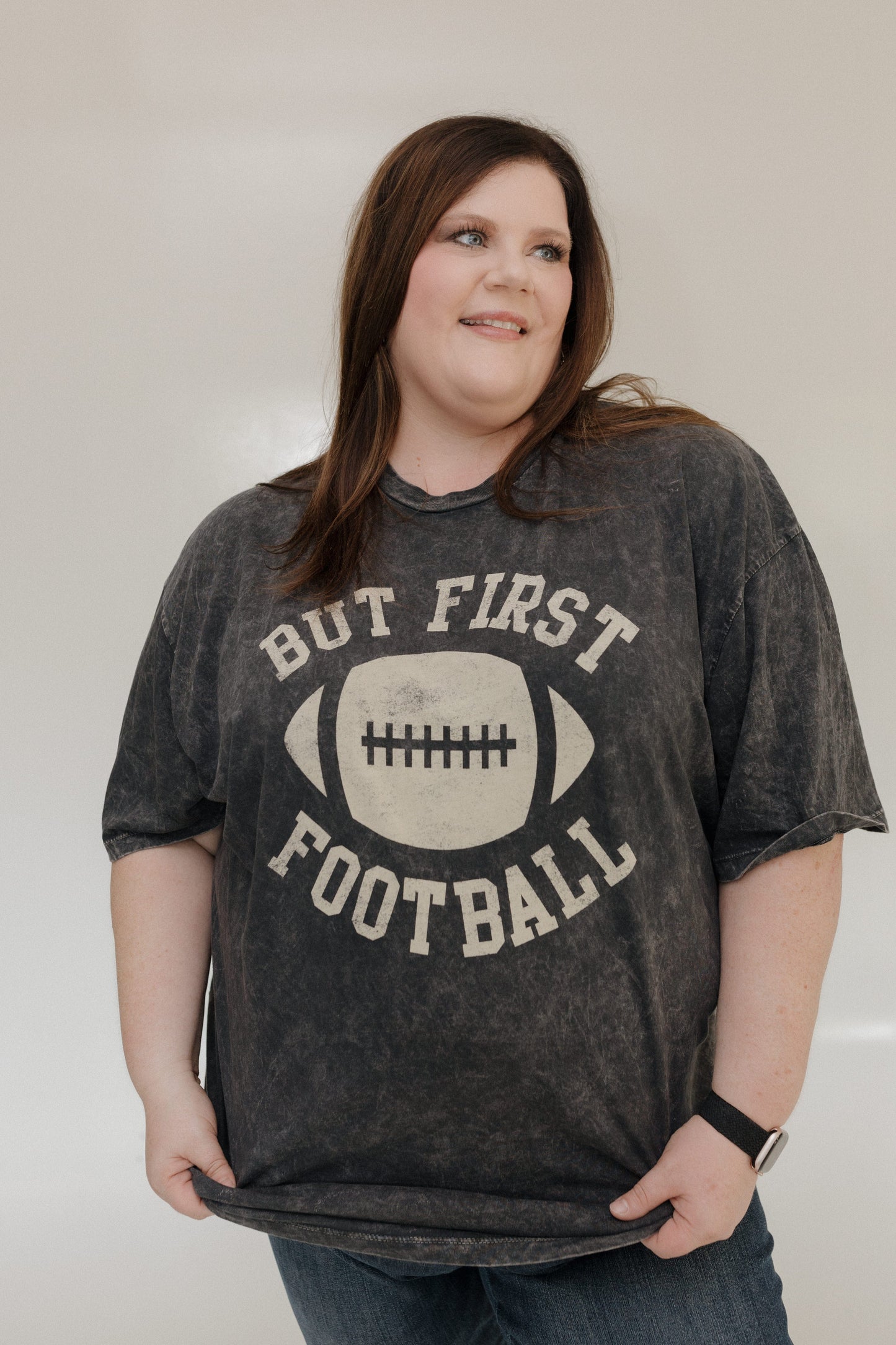 "BUT FIRST FOOTBALL" GRAPHIC TEE A person smiling while wearing a "But First Football" graphic tee from Love Marlow, made with super soft fabric and featuring a stylish mineral wash black finish. Love Marlow
