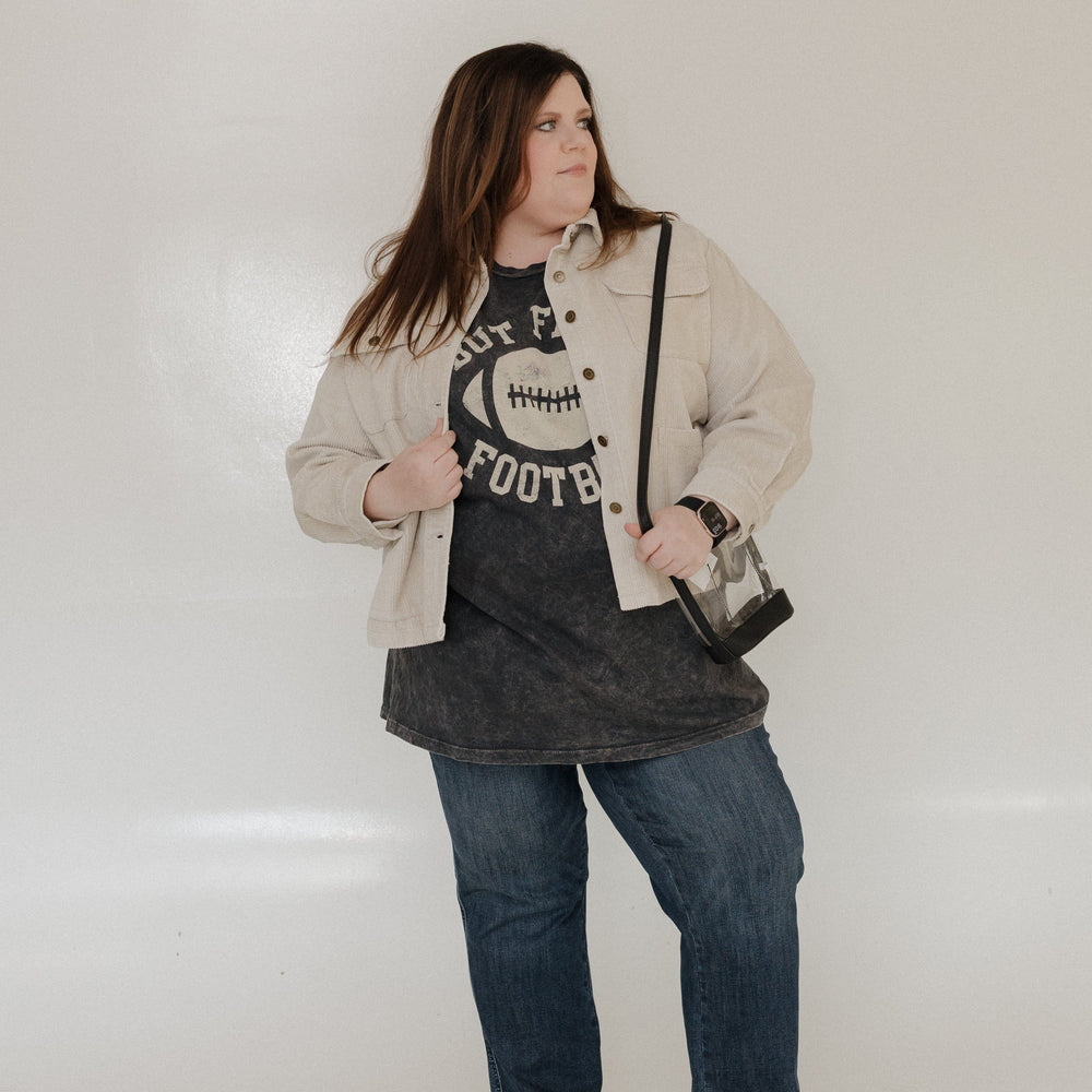 "BUT FIRST FOOTBALL" GRAPHIC TEE A person smiling while wearing a "But First Football" graphic tee from Love Marlow, made with super soft fabric and featuring a stylish mineral wash black finish. 