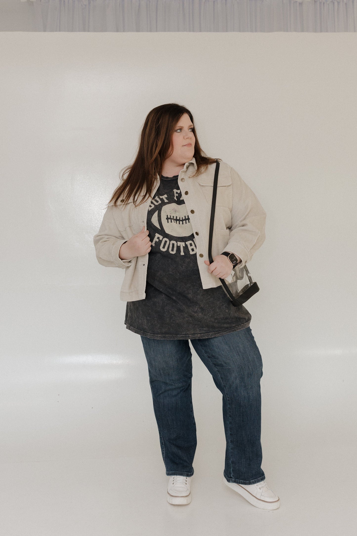 "BUT FIRST FOOTBALL" GRAPHIC TEE A person smiling while wearing a "But First Football" graphic tee from Love Marlow, made with super soft fabric and featuring a stylish mineral wash black finish. 