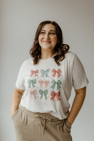 A person smiles while standing with hands in pockets. They are wearing a white t-shirt with a holiday bow pattern and beige pants, against a plain background.