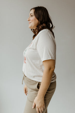 A person with long hair is smiling and looking to the right, wearing a white holiday graphic tee and beige pants against a neutral background.