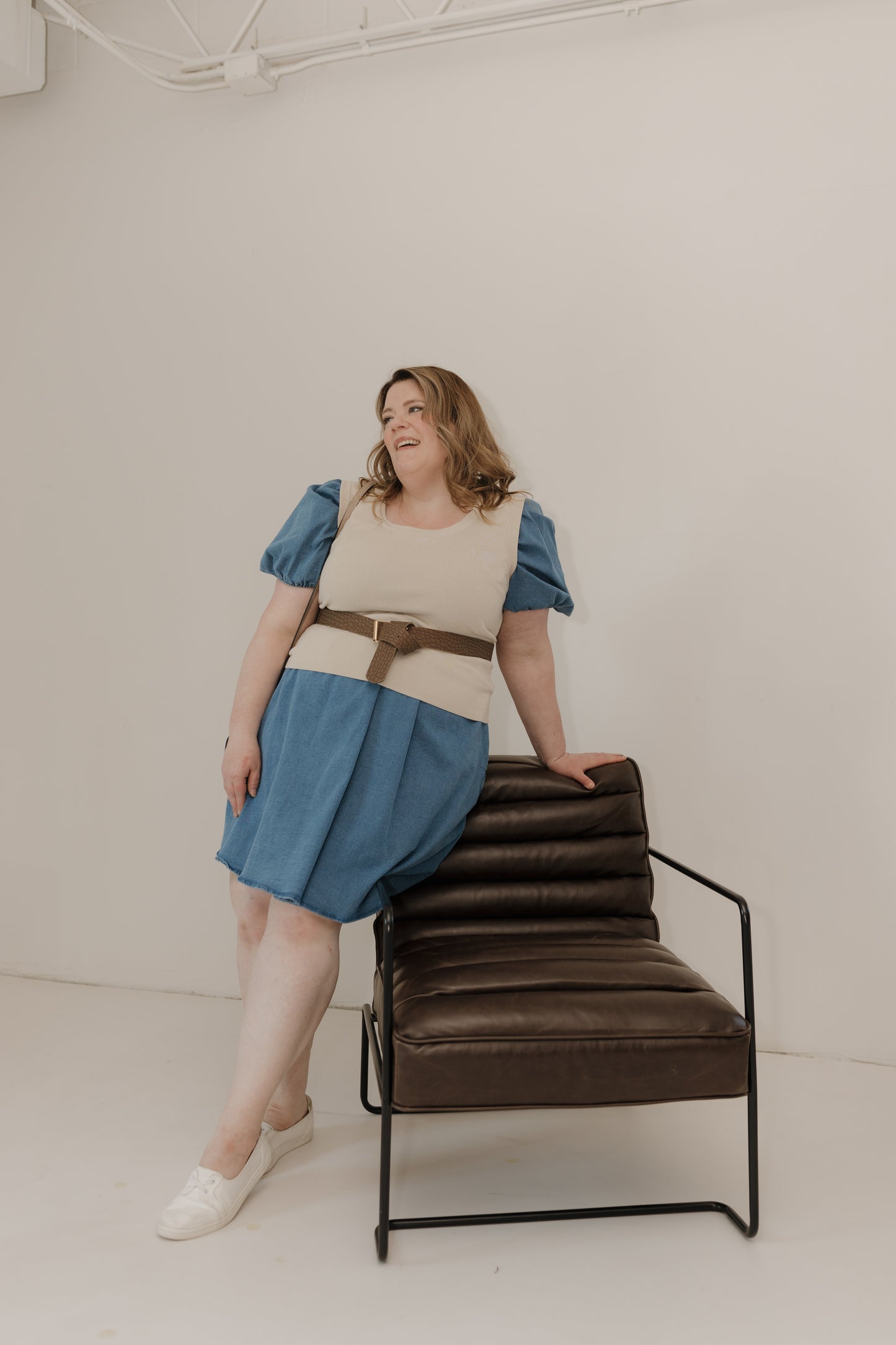 Woman in a blue dress and beige top standing next to a brown chair against a white background