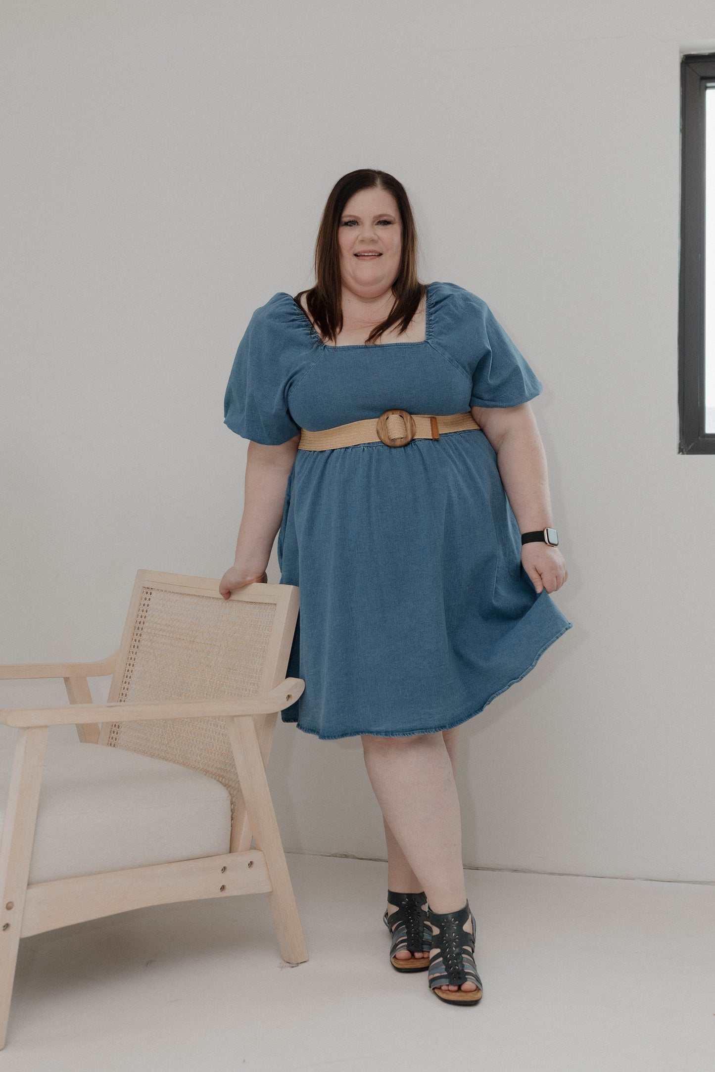Woman wearing a blue dress standing next to a wooden chair against a white wall.