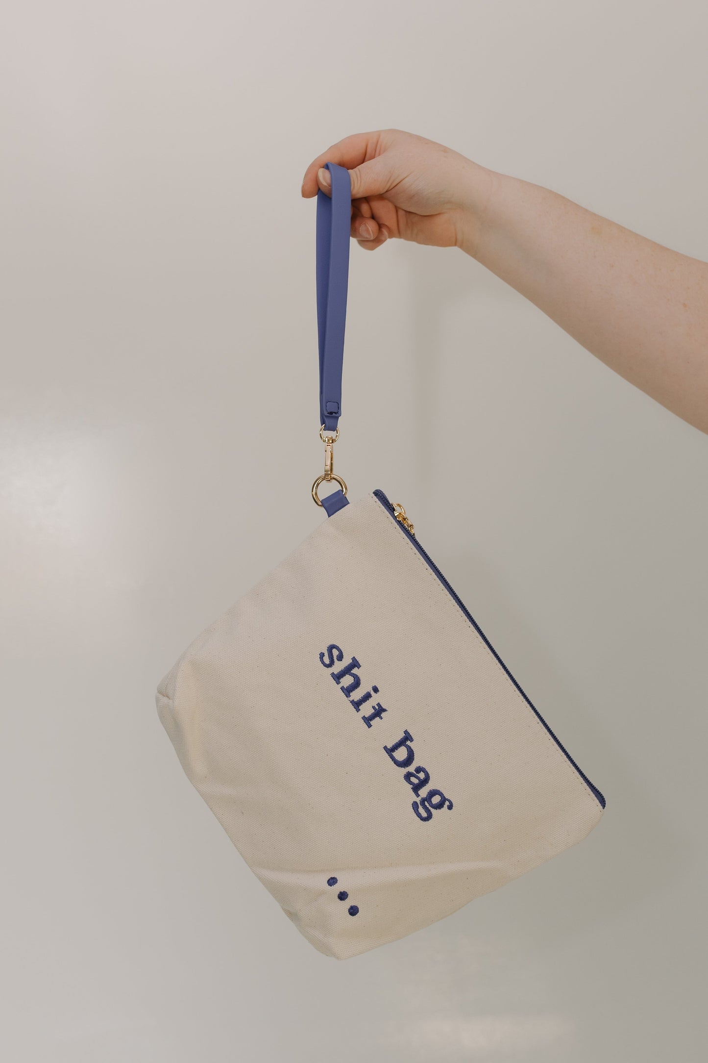 Beige pouch with 'shirt bag' text and blue handle held by a hand against a neutral background
