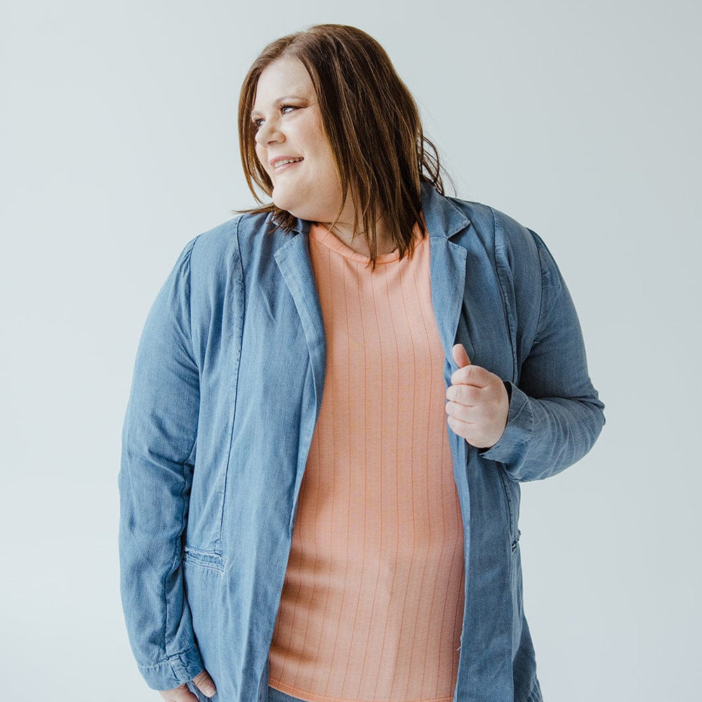 In front of a simple white backdrop, an individual wears the CHAMBRAY BOYFRIEND BLAZER by Dex, distinguished by its notched lapel, layered over a peach shirt.