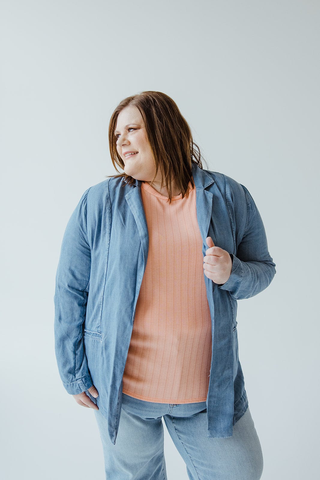 In front of a simple white backdrop, an individual wears the CHAMBRAY BOYFRIEND BLAZER by Dex, distinguished by its notched lapel, layered over a peach shirt.