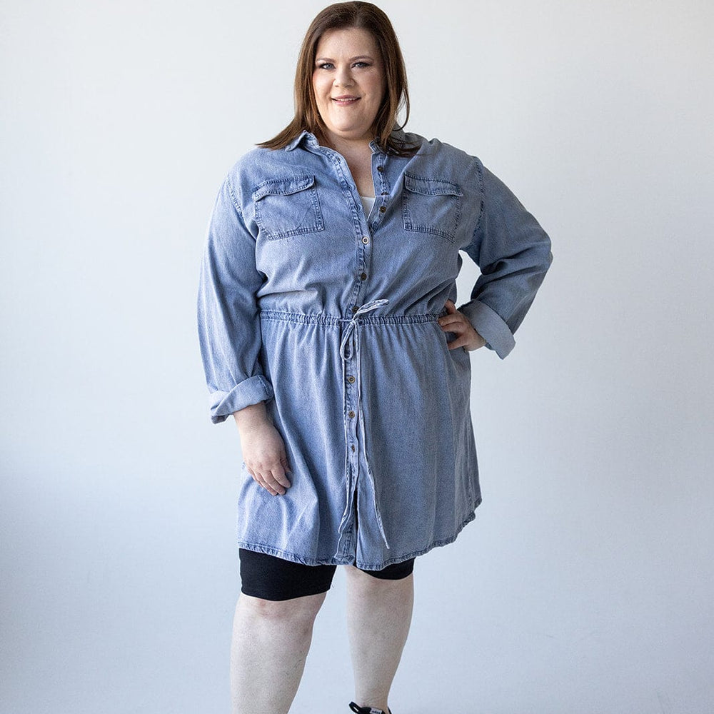 A person stands in a relaxed pose, wearing the CHAMBRAY SHIRTDRESS WITH DRAWSTRING WAIST by Love Marlow and black shoes, against a plain white background.