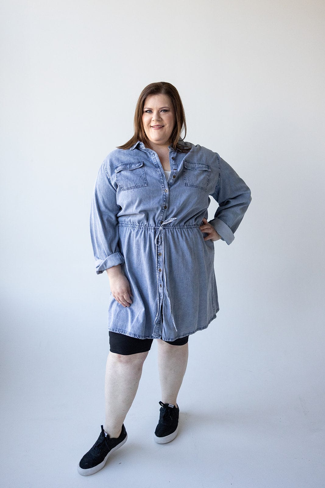 A person stands in a relaxed pose, wearing the CHAMBRAY SHIRTDRESS WITH DRAWSTRING WAIST by Love Marlow and black shoes, against a plain white background.