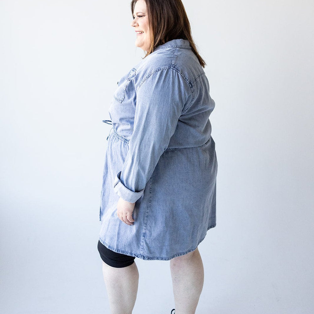 A woman wearing the Love Marlow CHAMBRAY SHIRTDRESS WITH DRAWSTRING WAIST and black sneakers stands sideways against a white background.