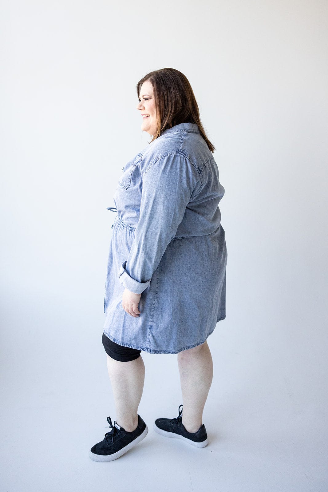 A woman wearing the Love Marlow CHAMBRAY SHIRTDRESS WITH DRAWSTRING WAIST and black sneakers stands sideways against a white background.