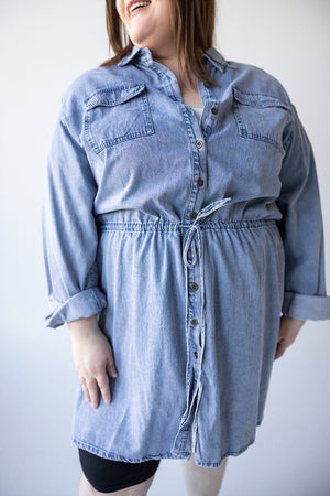 A person wearing the stylish Chambray Shirtdress with Drawstring Waist from Love Marlow, complete with rolled-up sleeves, smiles and looks to the side.