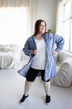 A person standing indoors near a window, smiling confidently with hands on hips, wearing the Chambray Shirtdress with Drawstring Waist by Love Marlow.