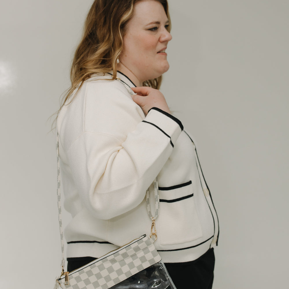 Woman wearing a white cardigan with black trim, holding a checkered handbag against a plain background