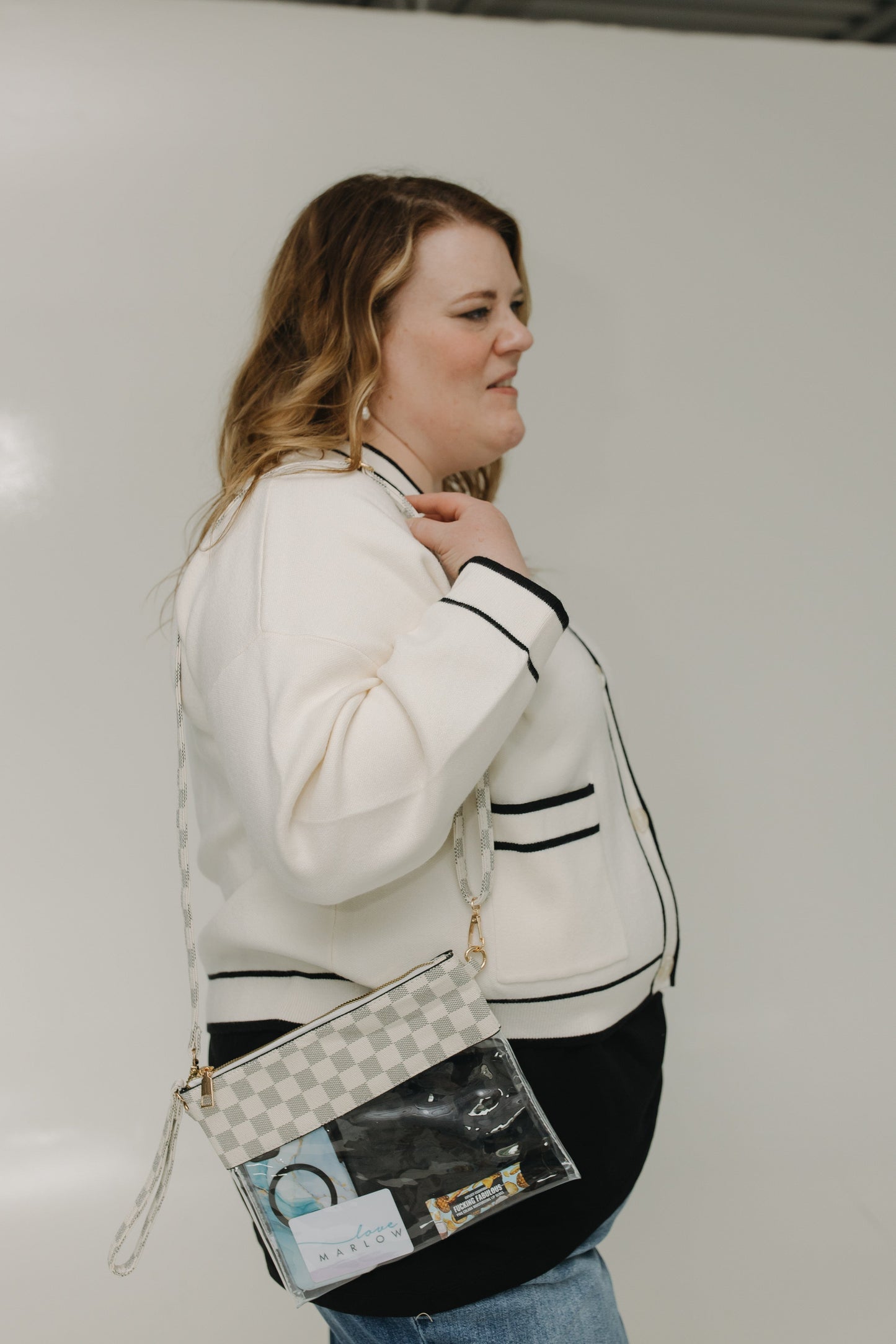 Woman wearing a white cardigan with black trim, holding a checkered handbag against a plain background