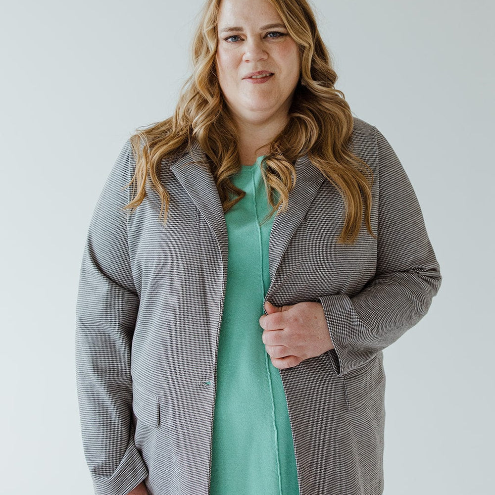 A woman with long hair, donning the luxurious CHECKED PATTERNED KNIT BLAZER by Love Marlow and a green top, stands facing forward against a plain background.