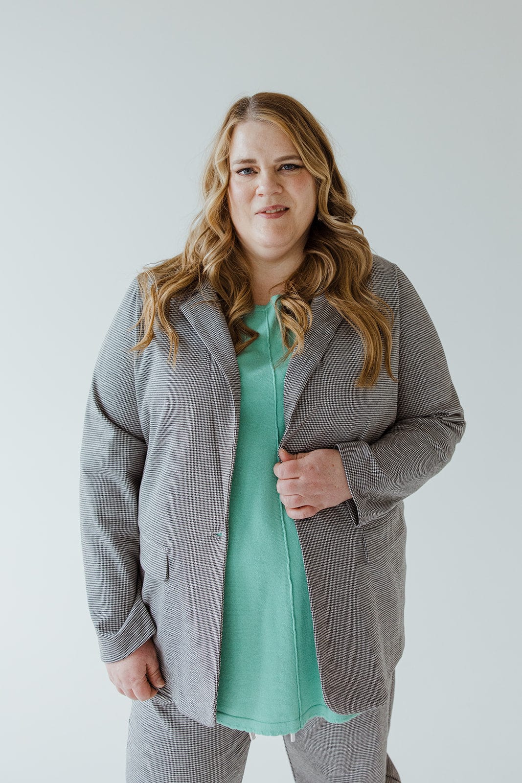 A woman with long hair, donning the luxurious CHECKED PATTERNED KNIT BLAZER by Love Marlow and a green top, stands facing forward against a plain background.