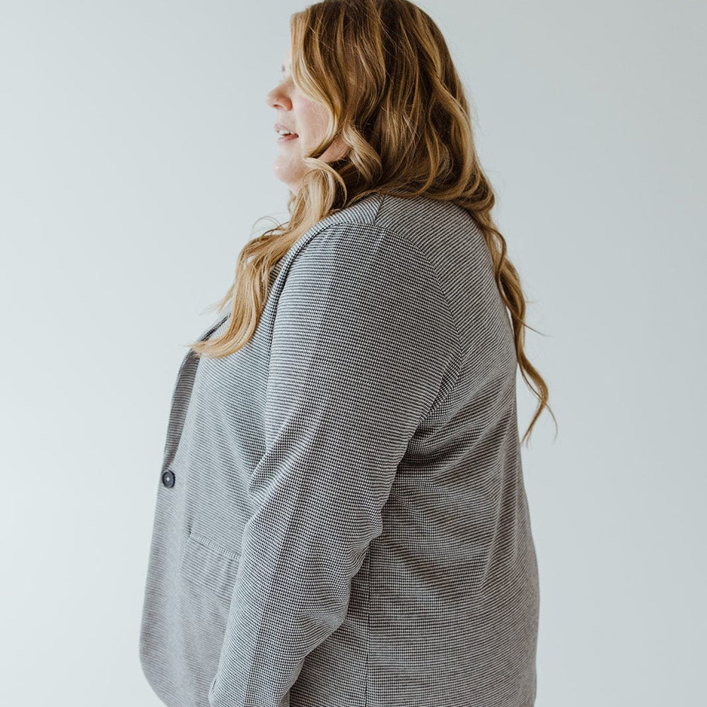 A person with long, wavy hair, wearing the luxurious CHECKED PATTERNED KNIT BLAZER by Love Marlow, stands in profile against a light grey background.