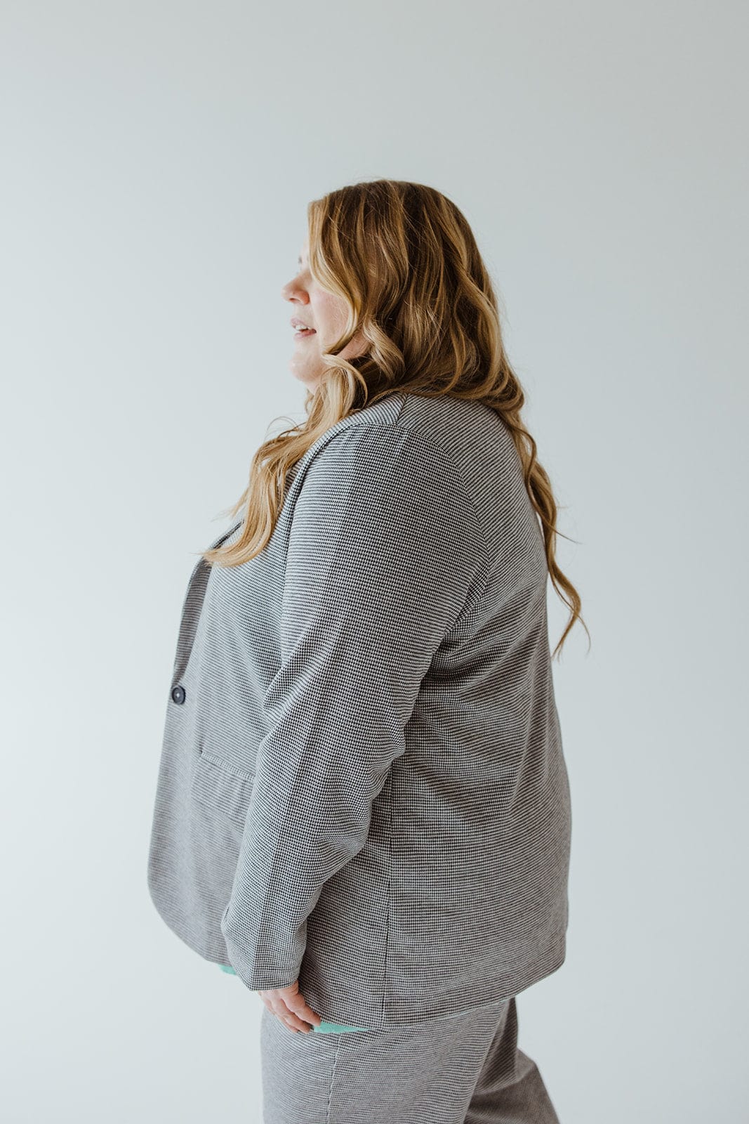 A person with long, wavy hair, wearing the luxurious CHECKED PATTERNED KNIT BLAZER by Love Marlow, stands in profile against a light grey background.