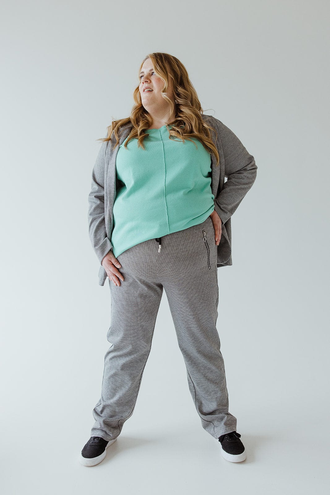 Person in gray tracksuit and mint green top stands with hands on hips, looking to the side, showcasing zipper detail pockets.