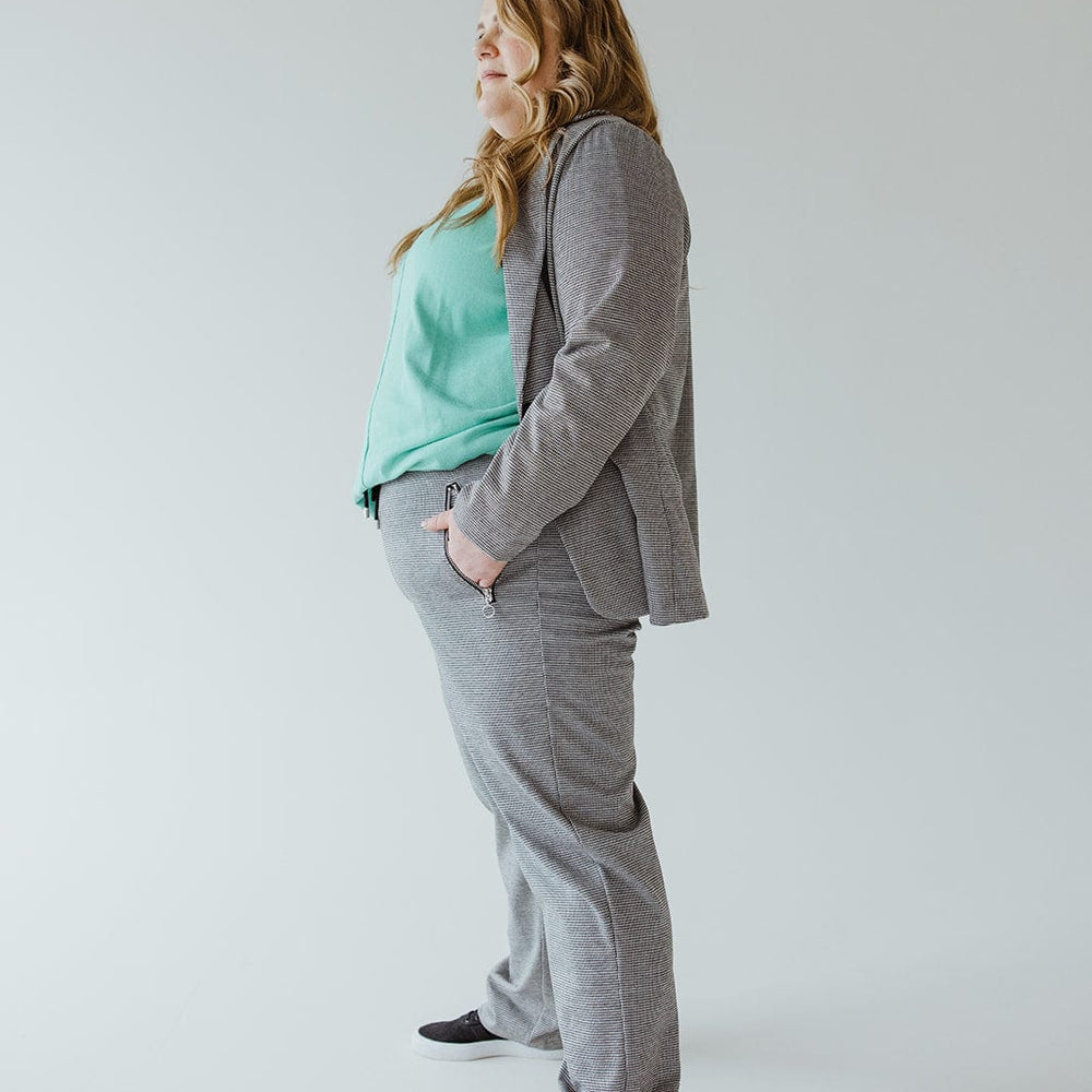 A person stands in profile wearing a gray suit and a light green top, complemented by checked patterned knit trousers. Their long hair rests over their shoulder, adding elegance against the plain background.