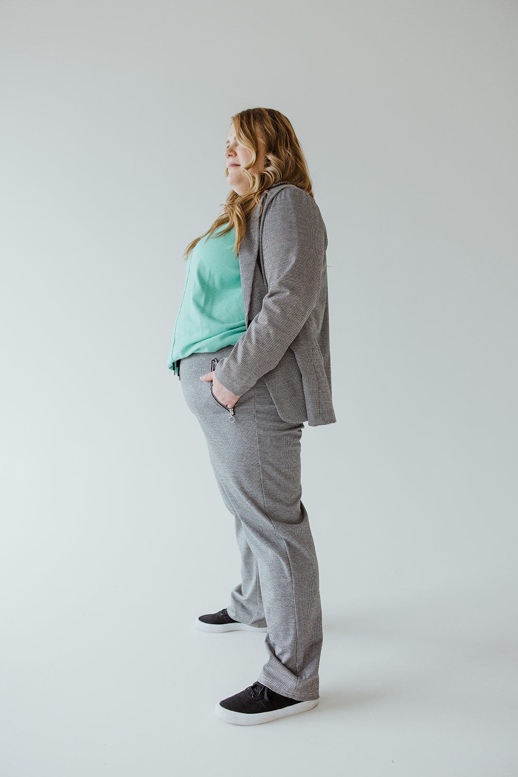 A person stands in profile wearing a gray suit and a light green top, complemented by checked patterned knit trousers. Their long hair rests over their shoulder, adding elegance against the plain background.