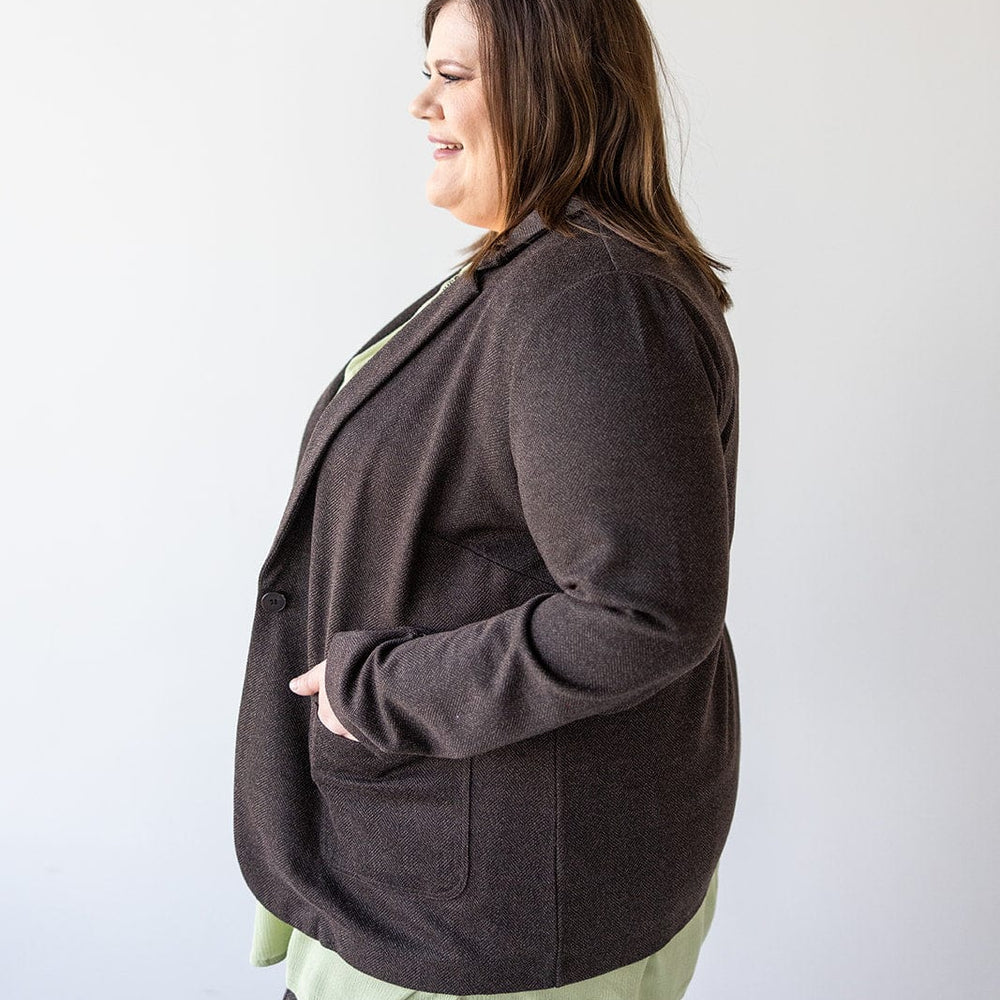 A person in a stylish CHOCOLATE BLAZER IN TWEED by Love Marlow, paired with a light green shirt, smiles while looking to the left against a plain background.