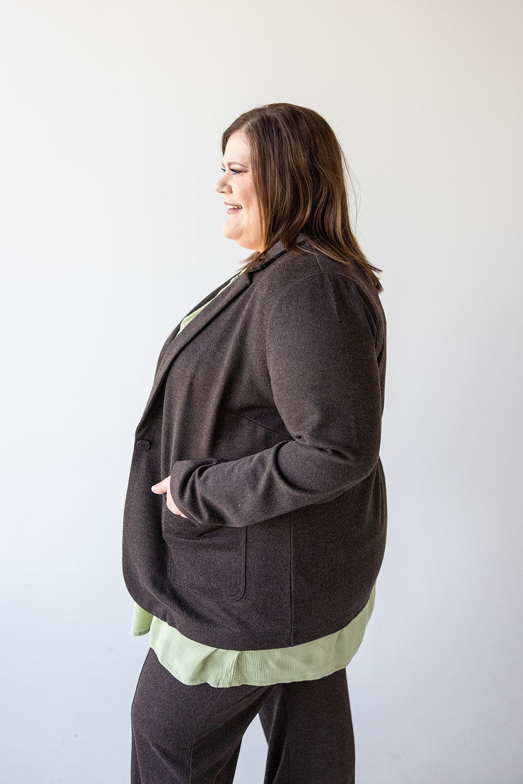 A person in a stylish CHOCOLATE BLAZER IN TWEED by Love Marlow, paired with a light green shirt, smiles while looking to the left against a plain background.