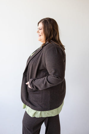 A person in a stylish CHOCOLATE BLAZER IN TWEED by Love Marlow, paired with a light green shirt, smiles while looking to the left against a plain background.