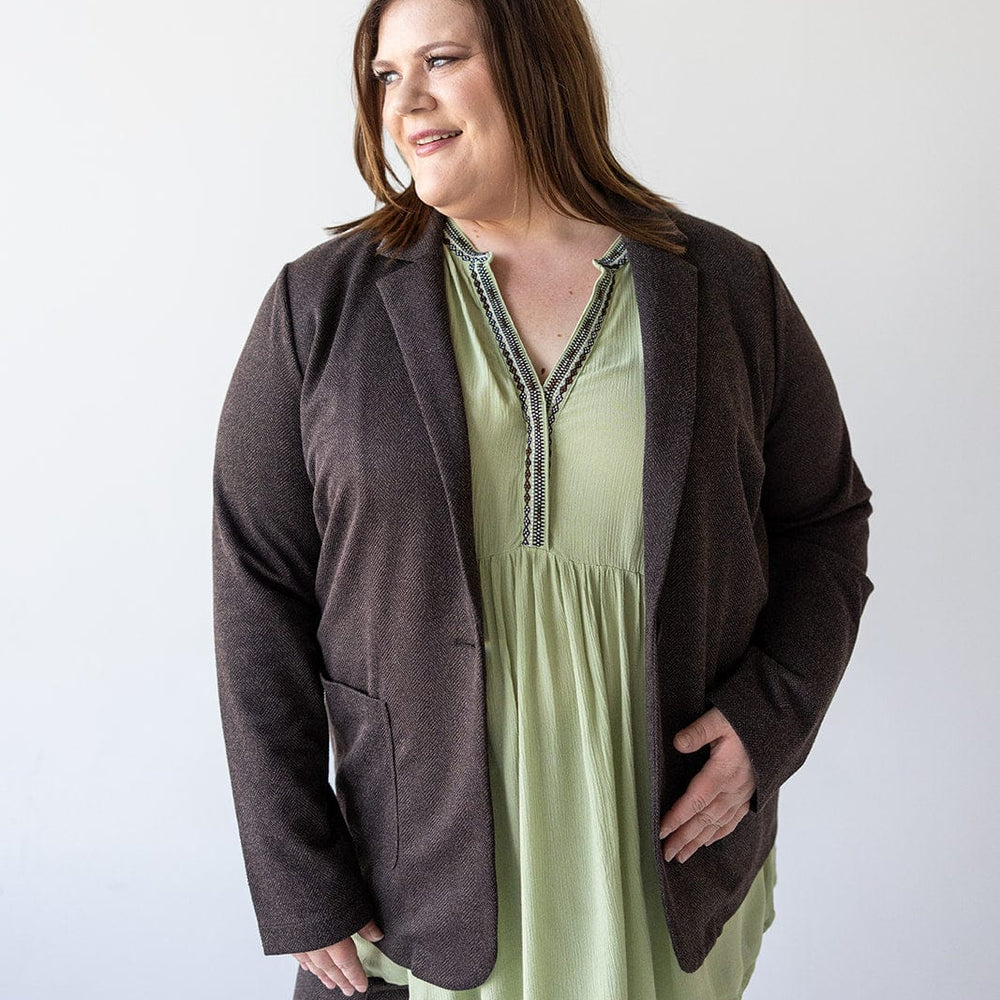 A person in a chic CHOCOLATE BLAZER IN TWEED by Love Marlow and a pastel green blouse stands against a plain white background, gazing sideways with a neutral expression.