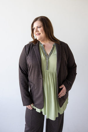 A person in a chic CHOCOLATE BLAZER IN TWEED by Love Marlow and a pastel green blouse stands against a plain white background, gazing sideways with a neutral expression.