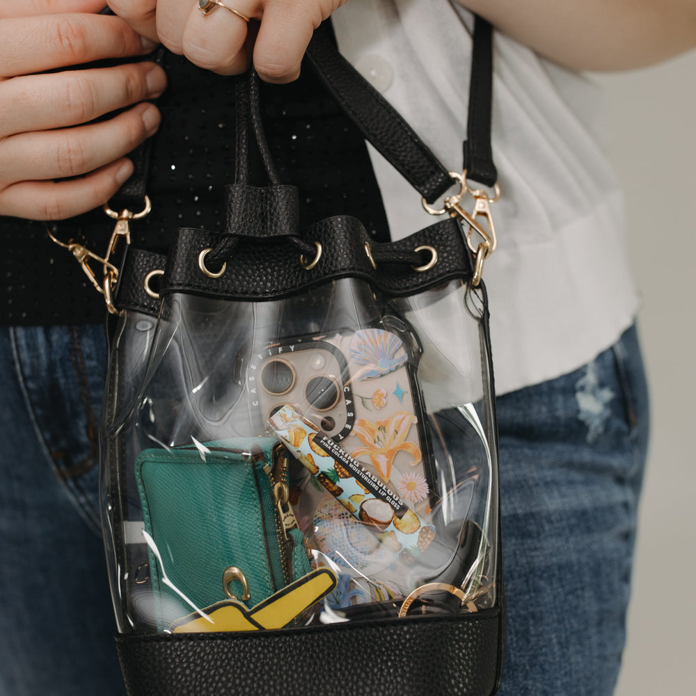 Clear handbag with black trim held by a person, showing various items inside.