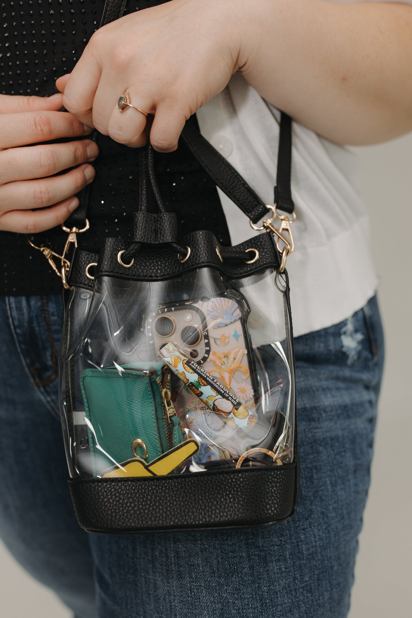 Clear handbag with black trim held by a person, showing various items inside.