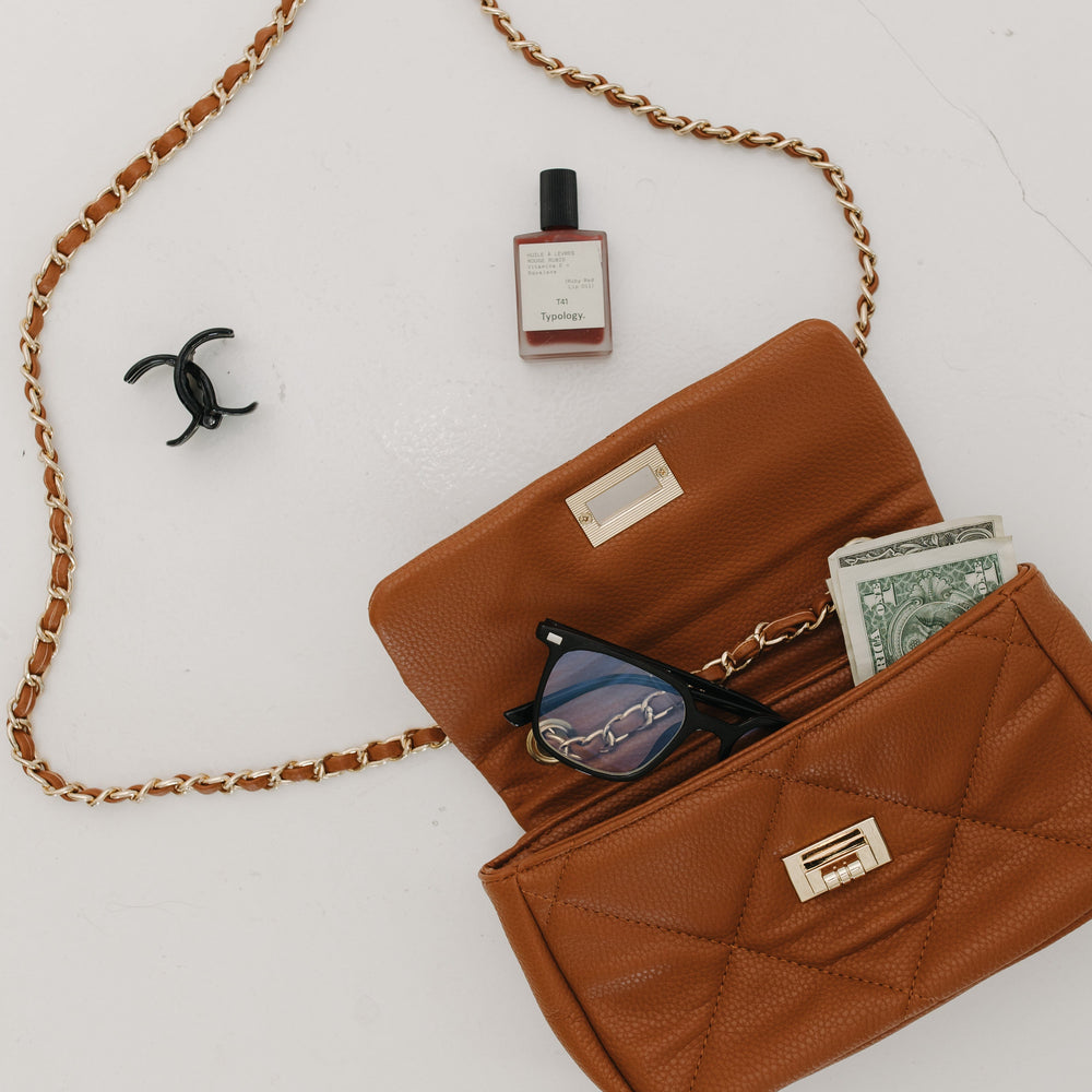 Brown handbag with chain strap, sunglasses, money, and a bottle on a white background