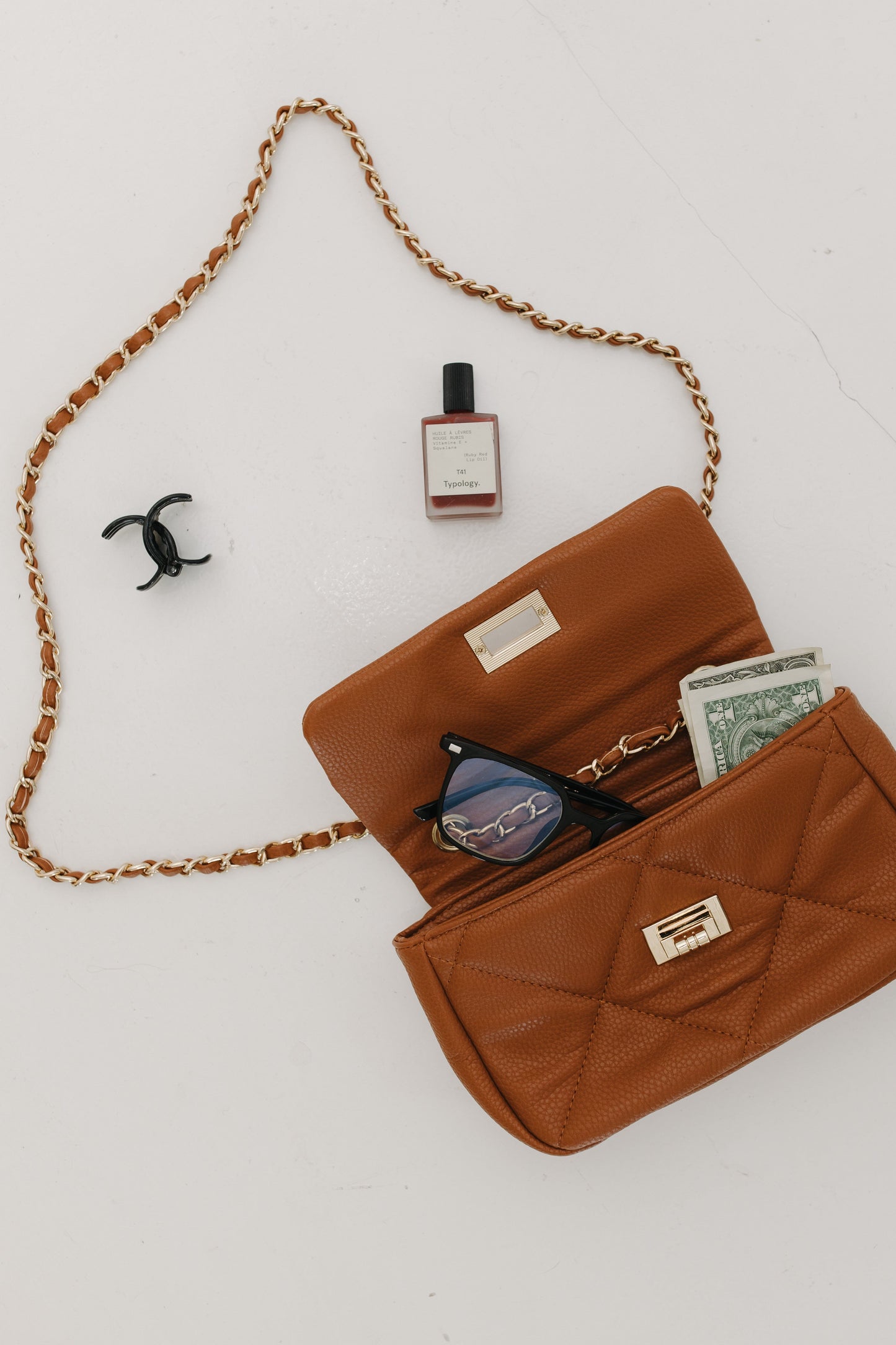 Brown handbag with chain strap, sunglasses, money, and a bottle on a white background